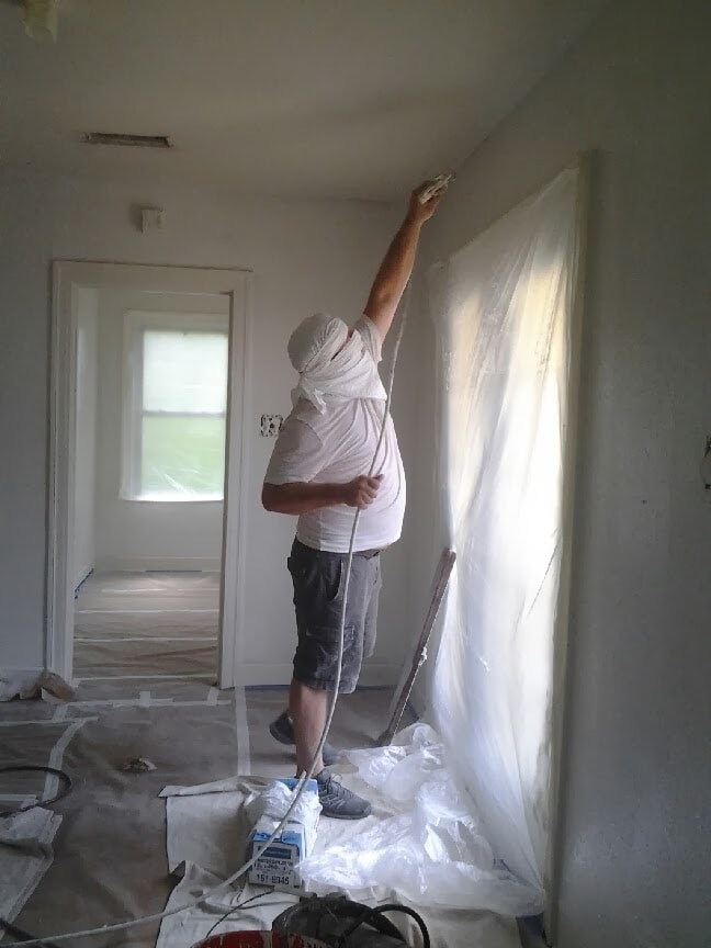 Painter spraying interior room walls with airless sprayer and plastic sheeting protection