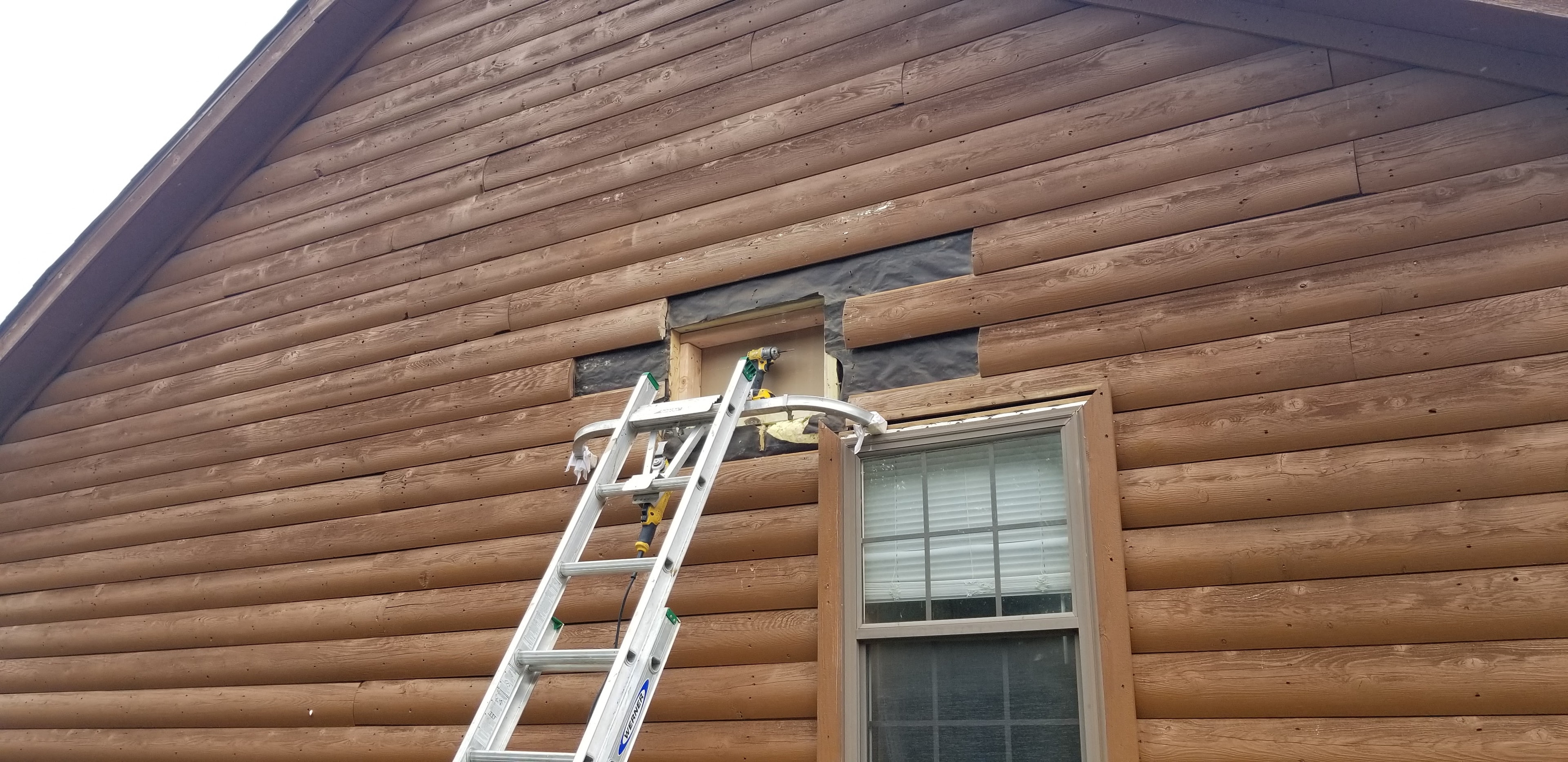 Log cabin siding repair in progress with ladder and drill at window opening on gable wall