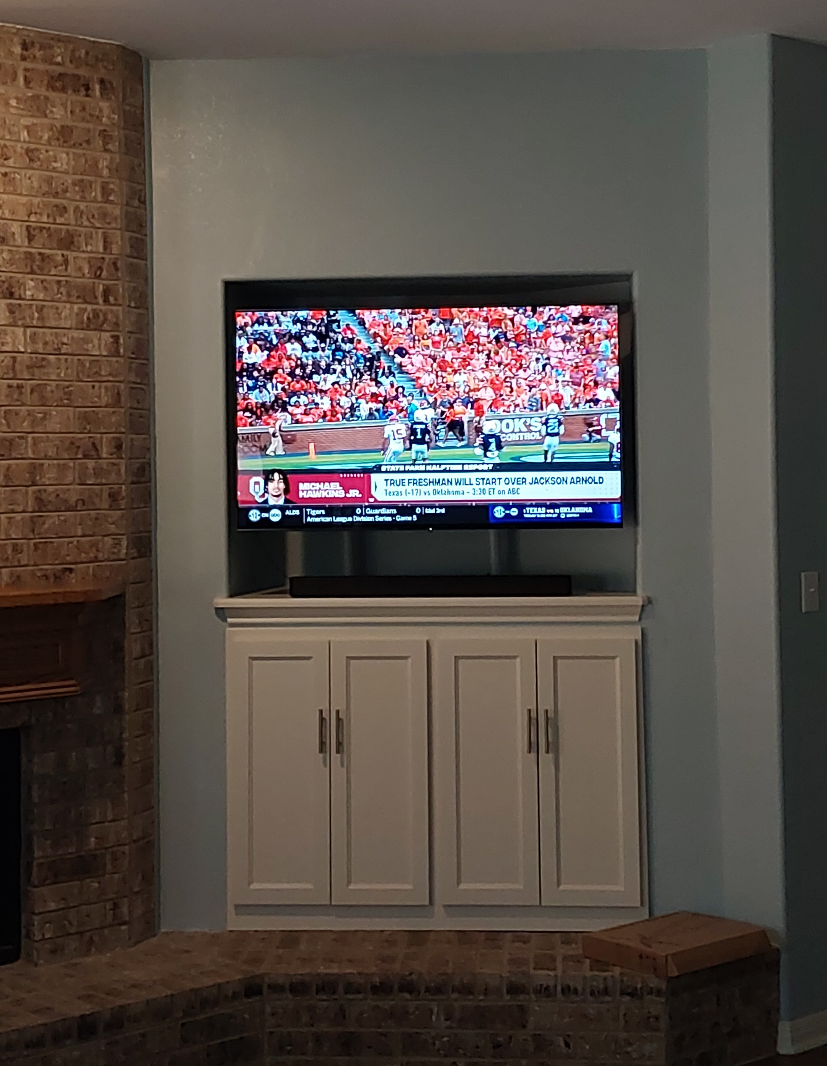 Finished TV entertainment center with white built-in cabinet below wall-mounted TV beside brick fireplace